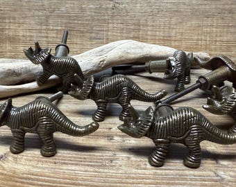 Op de afbeelding: Vijf antieke bronzen knoppen in de vorm van een dinosaurus met een gestructureerd oppervlak. De knoppen bevinden zich in verschillende poses, waaronder staand, lopend en rennend.