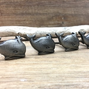 Peut inclure: Quatre boutons de meuble en forme de baleine en fonte, alignés. Les boutons sont de couleur gris foncé avec une finition texturée. Chaque baleine a une queue, une nageoire et un petit œil. Les boutons mesurent environ 5 cm de long.