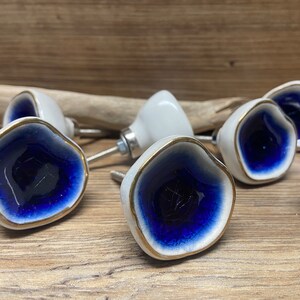 Puede incluir: Un conjunto de pomos de cerámica blanca con un diseño azul y dorado. Los pomos tienen una forma irregular con un centro azul oscuro y un borde dorado. El herraje está sobre un fondo de madera.
