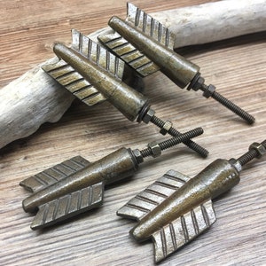 May include: Four arrow-shaped cabinet knobs made of antique bronze-toned metal. Each knob features a fletching design with textured grooves and includes a threaded metal bolt with a small nut for attachment to furniture or cabinetry doors.
