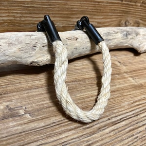 Peut inclure: Une poignée décorative en corde avec des fixations en métal noir. La corde blanc cassé est en forme de U. La poignée est posée sur un fond en bois et un morceau de bois flotté.