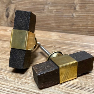 Pode incluir: Dois puxadores de armário de madeira castanha escura com uma faixa de metal cor de latão ao redor do centro. Os puxadores têm formato retangular e são montados em um parafuso prateado. O fundo é uma superfície de madeira castanha clara.