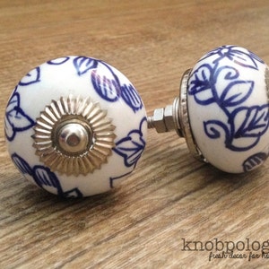 Può includere: Due pomelli per mobili in ceramica bianca con un motivo floreale blu. Ogni pomello ha una base in metallo argentato e un bullone per il fissaggio. I pomelli sono su una superficie di legno. Il testo "knobpologie" è visibile.