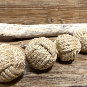 Peut inclure: Quatre boules décoratives en corde, chacune méticuleusement fabriquée avec de la corde de couleur naturelle. Les boules sont disposées en rangée sur une surface en bois, avec un morceau de bois flotté en arrière-plan. Sphères sur le thème nautique.