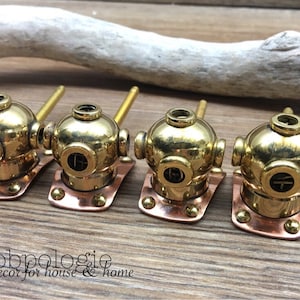 May include: Four brass and copper cabinet knobs in the shape of vintage diving helmets. Each knob features a round, brass helmet with a copper base and a brass tube. The knobs are arranged on a wooden surface.