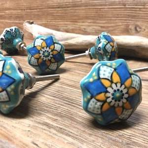 Könnte beinhalten: Eine Sammlung von Keramik-Schrankknöpfen mit floralem Design. Die Knöpfe sind türkis, mit blauen, gelben und weißen Blumenmustern. Jeder Knopf hat eine Metallschraube zur Befestigung. Die Knöpfe sind auf einer Holzoberfläche angeordnet.