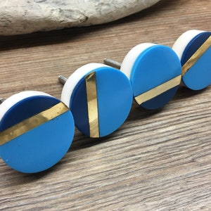May include: Four decorative cabinet knobs. Each knob has a white base, a section of navy blue, a section of light blue, and a gold-coloured diagonal stripe. The knobs are round and have a metal screw for installation.