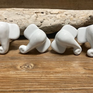 Peut inclure: Quatre boutons de meuble blancs en céramique en forme d'éléphant. Les boutons sont disposés sur une surface en bois, avec un morceau de bois flotté en arrière-plan. Chaque bouton présente une trompe détaillée, des oreilles et une finition lisse et brillante.