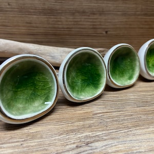May include: Four ceramic cabinet knobs with a white exterior, a gold rim, and a green interior. The knobs have a unique, organic shape and are arranged in a row. The knobs are on a wooden surface.