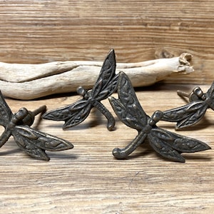 May include: Four antique bronze dragonfly drawer pulls with intricate details. The pulls are in the shape of a dragonfly with its wings spread out. They are perfect for adding a touch of nature to any home decor.