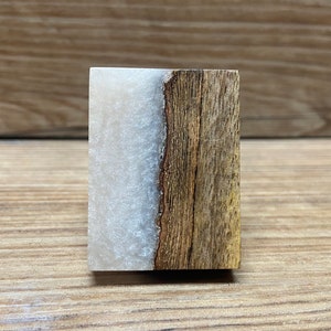 Puede incluir: Un bloque rectangular compuesto por dos secciones distintas unidas verticalmente. Un lado presenta madera marrón natural con una veta visible y un borde texturizado, mientras que el otro lado consiste en un material de resina blanca semitranslúcida.