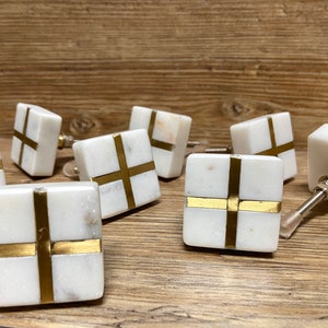 Könnte beinhalten: Eine Sammlung quadratischer Schrankknöpfe aus weißem Marmor mit goldfarbenen Kreuzakzenten. Die Knöpfe haben eine polierte Oberfläche und sind auf einer Holzoberfläche angeordnet, um ihr Design und ihre Textur hervorzuheben.