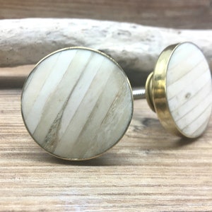 Puede incluir: Dos tiradores redondos para armario con un diseño a rayas crema y beige. Los tiradores tienen una base de metal dorado y un tornillo para la instalación. Los tiradores están sobre un fondo de madera clara.