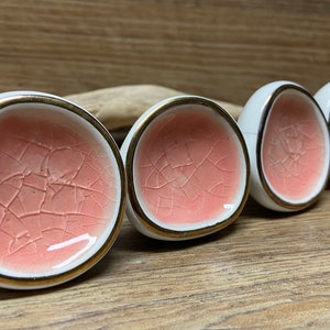 May include: Four white ceramic cabinet knobs with a cracked coral-colored interior and gold trim. The knobs are arranged in a row, showcasing their unique design and texture. The background is a wooden surface.