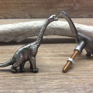 Op de afbeelding: Twee zilveren dinosaurus-ladegrepen. Eén dinosaurus staat rechtop met zijn nek uitgestrekt. De andere dinosaurus ligt met zijn staart opgerold. Beide dinosaurussen hebben een getextureerd oppervlak.