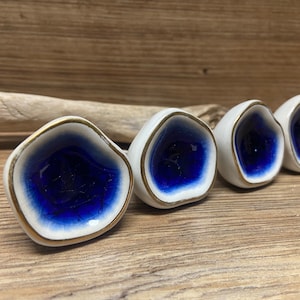 Puede incluir: Cuatro pomos de cerámica blanca con centro azul oscuro y borde dorado. Los pomos tienen una forma orgánica e irregular, dispuestos en fila. El fondo es una superficie de madera.
