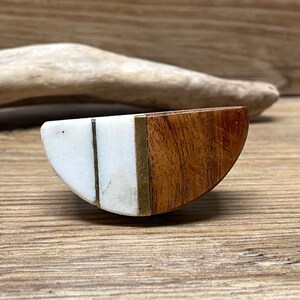May include: A half-moon shaped decorative cabinet knob. The knob is made of white marble, wood, and brass. The wood is a warm brown color, and the marble is white with subtle veining. The brass accents add a touch of elegance.