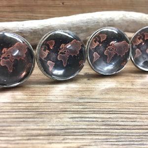 Può includere: Quattro pomelli rotondi per mobili con un design a mappa del mondo. I pomelli hanno uno sfondo scuro con una mappa del mondo color rame. I pomelli sono inseriti in una base metallica color argento. I pomelli sono esposti su una superficie di legno.