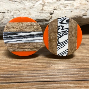 May include: A pair of circular stud earrings. Each has an orange base, with a central design. One features a wood grain and a black and white stripe. The other has wood grain and a black and white marbled stripe.
