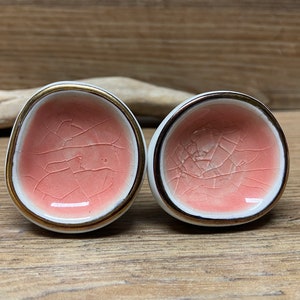 May include: Two ceramic knobs with a crackle glaze in a soft coral color. The knobs have a white base with a gold-colored rim. The knobs are likely for furniture or decorative use.