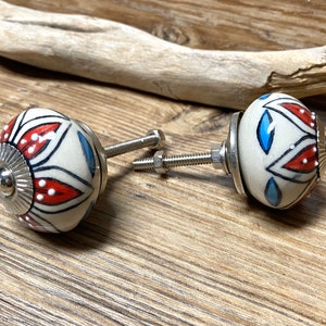 May include: Two decorative ceramic cabinet knobs. Each knob is white with a floral design in red, blue, and black. The knobs have silver metal hardware and are set against a wooden background.