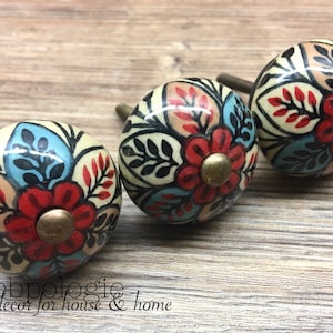 May include: Three decorative ceramic cabinet knobs with floral designs. Each knob features a cream-colored base with red, blue, and black floral patterns. The knobs have brass-colored hardware and are arranged on a wooden surface. The text "Knobpologie fresh decor for house & home" is visible.