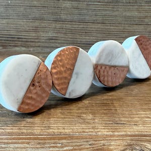 May include: Four circular cabinet knobs with a split design. One half is white, the other is hammered copper. The knobs are arranged in a row on a wooden surface. These decorative knobs can be used for furniture.