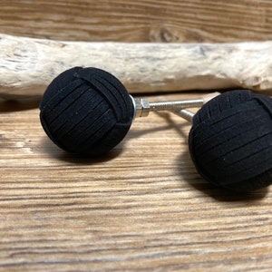 Può includere: Due pomelli neri e sferici per armadi con un aspetto strutturato e intrecciato. Ogni pomello è attaccato a una vite in metallo argentato con un dado. I pomelli sono esposti su una superficie di legno con un pezzo di legno alla deriva sullo sfondo.