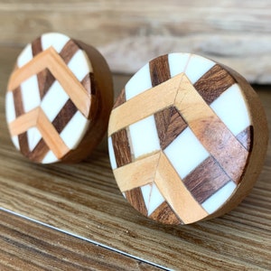 May include: Two round wooden knobs with a geometric pattern of white, brown, and light brown wood. The knobs have a smooth, polished finish.