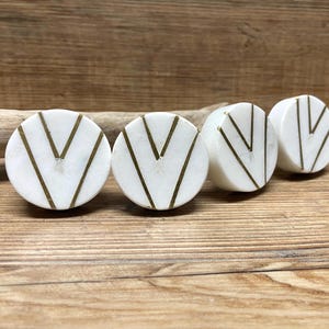 May include: Four white marble cabinet knobs with gold-colored chevron designs. The knobs are round and rest on a wooden surface. The knobs are a decorative element for furniture.