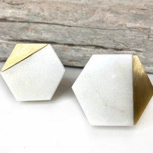 May include: Two hexagonal cabinet knobs featuring a white stone base with contrasting brushed gold metal accents. One knob has a gold triangular corner, while the other features a gold side panel, both showcasing a geometric design.