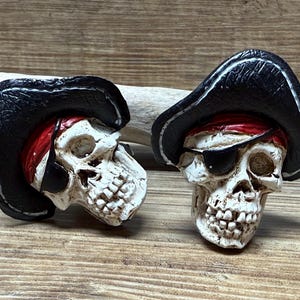 Puede incluir: Dos figuritas de calavera de pirata blancas con sombreros de pirata negros y bandanas rojas. Una calavera tiene un parche en el ojo.