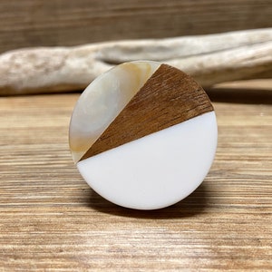 Könnte beinhalten: Ein runder Schrankknopf mit einem einzigartigen Design. Der Knopf ist in drei Bereiche unterteilt: weiß, Holzmaserung und ein perlmuttartiges Material. Der Knopf befindet sich auf einer Holzoberfläche.