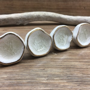 May include: Four small, white ceramic bowls with irregular edges and gold rims. The bowls have a crackle glaze interior and are arranged in a row on a wooden surface. A piece of driftwood is in the background.