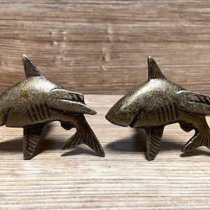 Op de afbeelding: Twee bronskleurige kastknoppen in de vorm van een haai. Elke knop heeft een gedetailleerd haaienontwerp met een rugvin, kieuwen en staartvin. De knoppen zijn gemonteerd op een houten oppervlak.