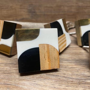 May include: Set of four square cabinet knobs with a geometric design. The knobs are made of wood, brass, and black resin. Each knob features a different combination of materials and colors.