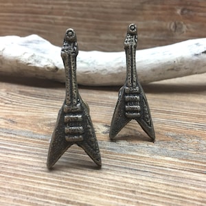 May include: Two bronze-coloured miniature electric guitar sculptures. The guitars have a V-shaped body and a long neck with detailed fretwork. They are displayed on a wooden surface with a piece of driftwood in the background.