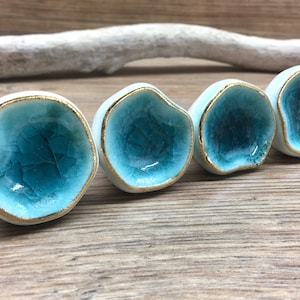 May include: Four ceramic cabinet knobs with a unique design. Each knob features a white exterior, a turquoise interior, and a gold-colored rim. The knobs have an organic, irregular shape, adding a touch of artistic flair to any furniture piece.