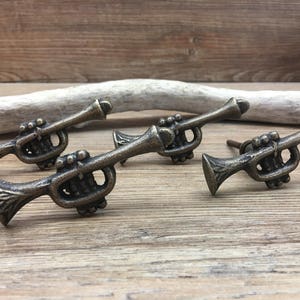 May include: Four bronze-colored trumpet-shaped cabinet pulls. The pulls are arranged on a wooden surface, with a piece of driftwood in the background. Each pull has intricate details, including valves and a bell.