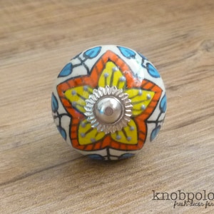 Könnte beinhalten: Dekorativer Keramik-Schrankknopf mit floralem Design. Der Knopf hat eine zentrale gelbe Blume mit orangefarbenen und blauen Akzenten und eine silberne Metallbasis. Der Text "knobpologie" ist sichtbar.