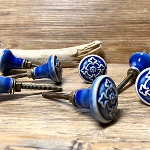 Può includere: Una collezione di pomelli per mobili in ceramica blu e bianca con un motivo floreale. I pomelli sono montati su viti e rondelle metalliche, su uno sfondo di legno. I pomelli sono di un blu brillante e lucido.