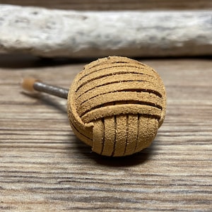 Może przedstawiać: Jasnobrązowy skórzany uchwyt o kulistym kształcie, z poziomymi paskami owiniętymi wokół centralnej kuli. Uchwyt jest przymocowany do metalowego pręta z małą, jasno zabarwioną końcówką. Tło to drewniana powierzchnia.
