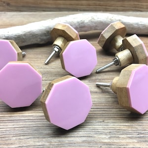 May include: A set of eight cabinet knobs. Each knob has a light pink, octagonal face and a natural wood base. The knobs are attached to silver screws. These knobs are designed for furniture and home decor.