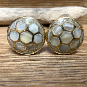 May include: A pair of cabinet knobs. Each knob has a gold-coloured metal rim and a surface of white and cream-coloured hexagonal mother-of-pearl tiles. The knobs are set against a wooden background.