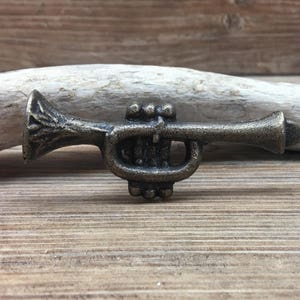 May include: A miniature brass trumpet figurine with intricate detailing. The small instrument is a muted gold color and rests on a wooden surface with a piece of driftwood in the background. The trumpet's bell and valves are clearly visible.