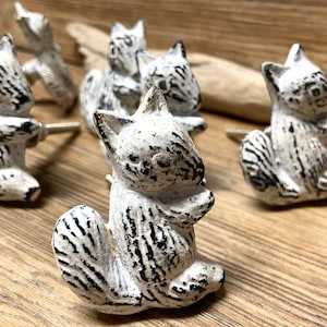 Puede incluir: Un conjunto de pomos de armario blancos con forma de ardilla y detalles en negro. Los pomos están hechos de un material envejecido, lo que les da un aspecto rústico. Las ardillas están sentadas con las patas juntas.