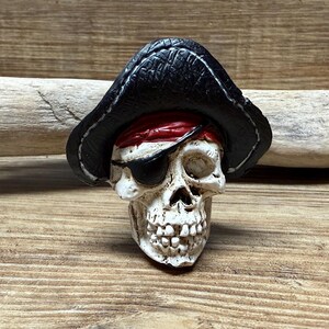 May include: A white pirate skull with a black leather pirate hat and a red bandana. The skull has one eye patch and is missing the other eye.