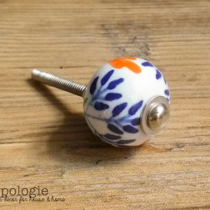 May include: A white ceramic cabinet knob with a floral design in orange and blue. The knob has a silver metal base and screw. The knob is on a wooden surface.