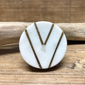 May include: A round, white marble cabinet knob with a brass chevron design. The knob is set against a wooden background, showcasing its natural stone texture and metallic accents. This decorative hardware adds a touch of elegance.
