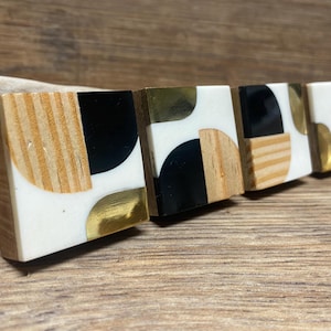 May include: A set of five decorative wooden blocks with a geometric design. Each block features a combination of wood grain, white resin, black semi-circles, and gold-colored metal accents. The blocks are arranged in a row on a wooden surface.